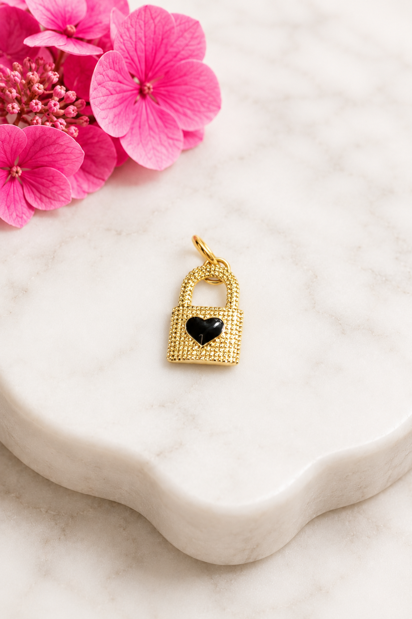 Gold lock-shaped pendant with a black heart on a marble surface with pink flowers.