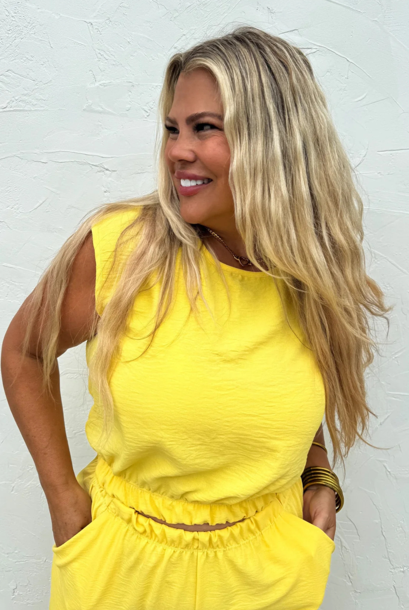 A woman with long blonde hair wearing a bright yellow sleeveless top and matching yellow bottoms, standing against a textured white wall.