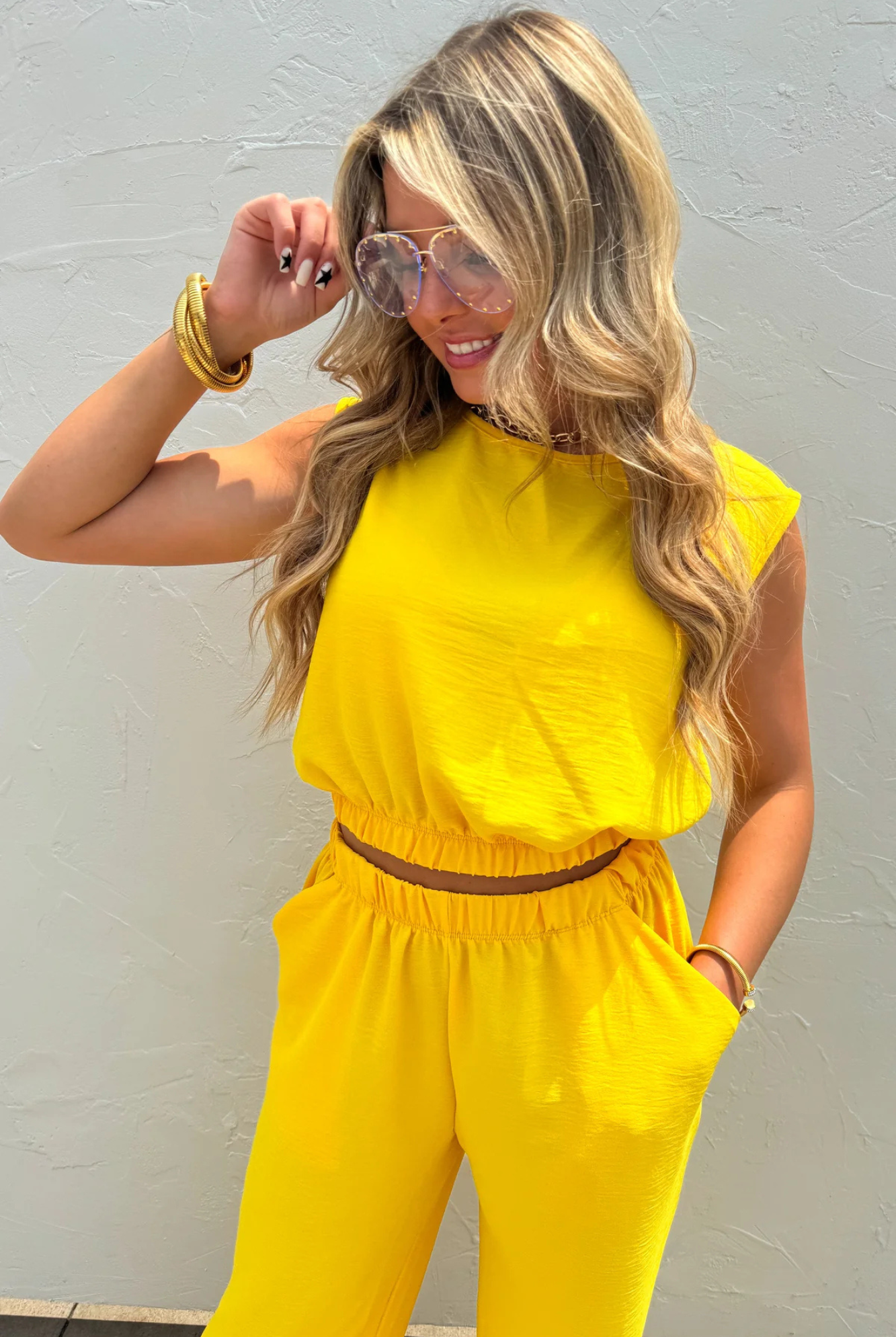 A woman wearing a bright yellow sleeveless jumpsuit with a thin brown belt at the waist, styled with gold bracelets and sunglasses, posing against a light gray textured wall.