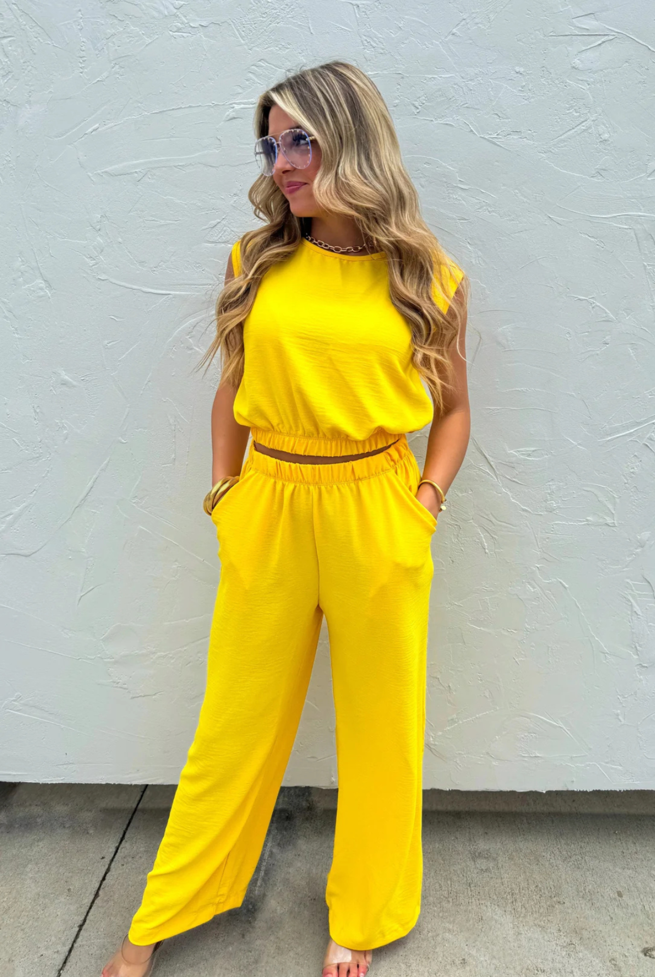 A woman modeling a bright yellow two-piece jumpsuit with a sleeveless top and wide-leg pants, standing against a textured white wall.