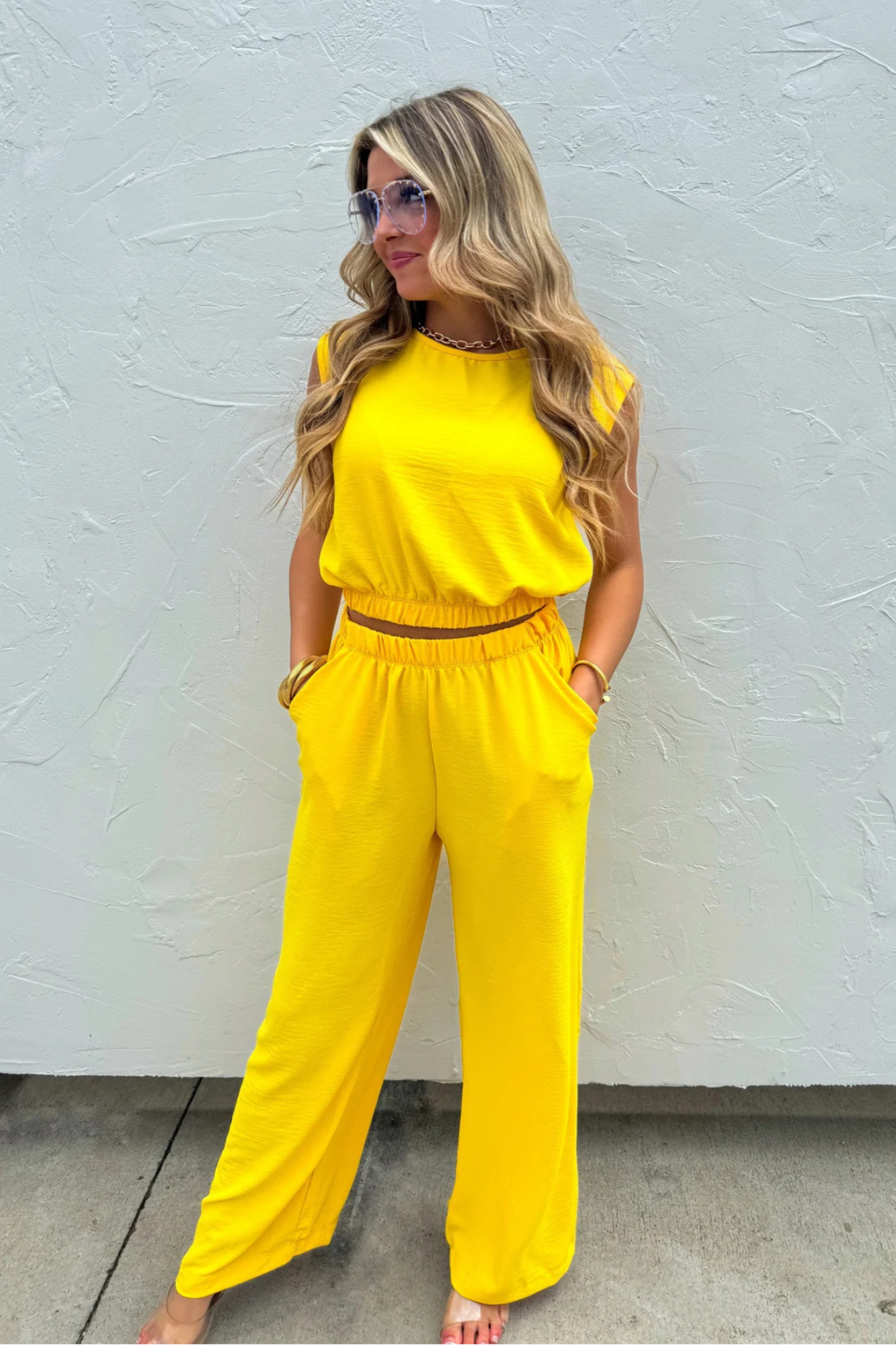 A woman modeling a bright yellow two-piece jumpsuit with a sleeveless top and wide-leg pants, standing against a textured white wall.