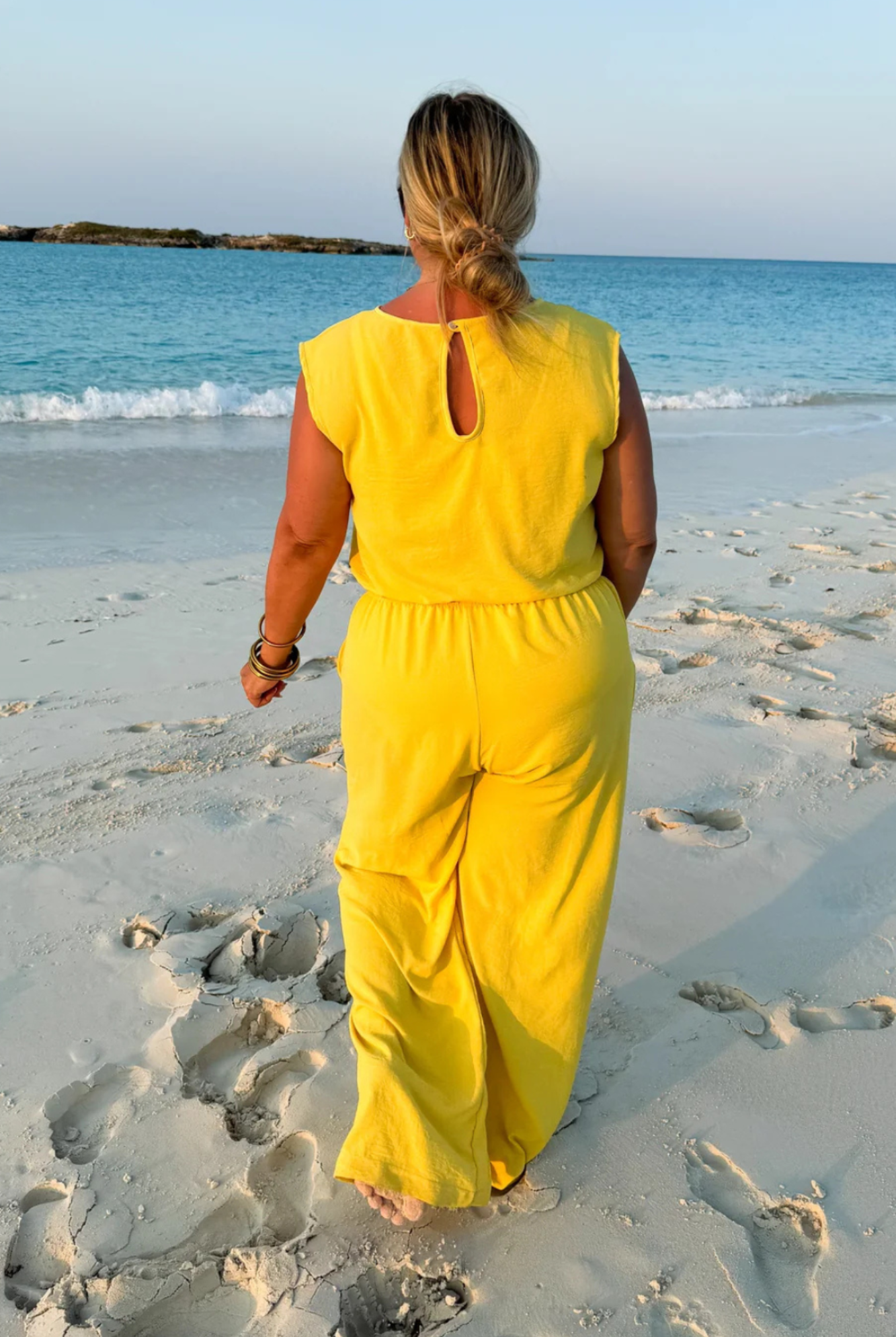 A woman seen from behind walking on a sandy beach toward the ocean, wearing a bright yellow sleeveless jumpsuit.