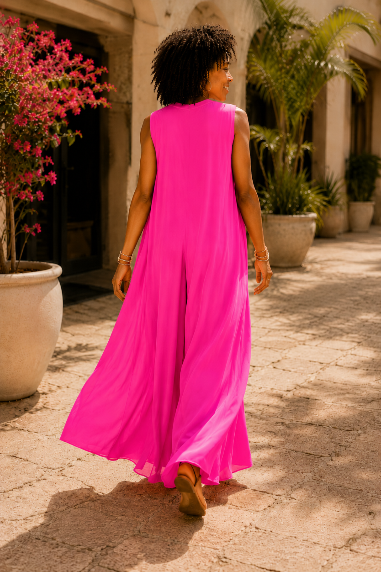 A woman wearing a bright hot pink sleeveless maxi dress walks away in a sunlit outdoor courtyard with potted plants and stone paving.
