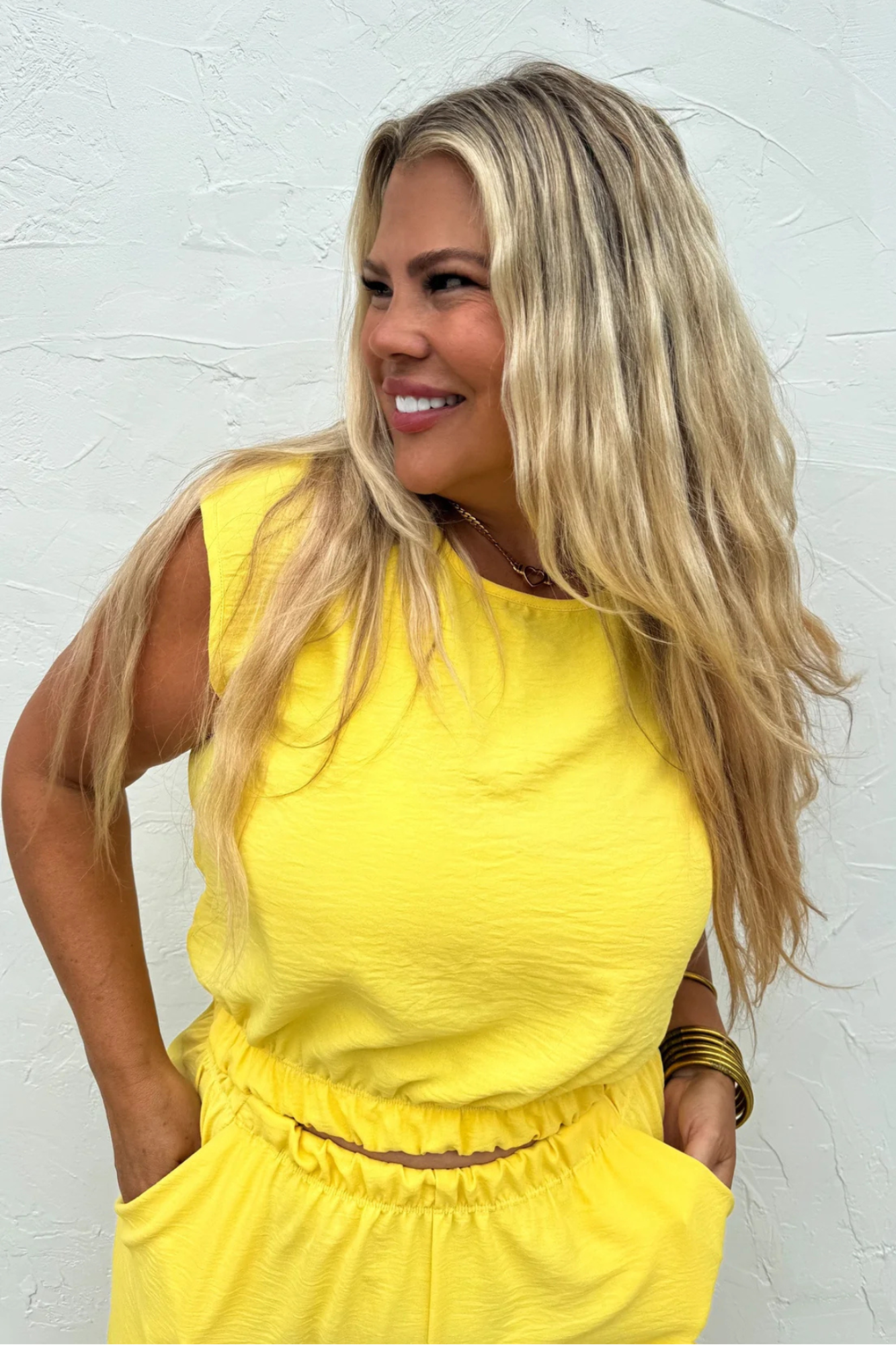 A woman with long blonde hair wearing a bright yellow sleeveless top and matching yellow bottoms, standing against a textured white wall.