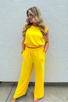 A woman modeling a bright yellow two-piece jumpsuit with a sleeveless top and wide-leg pants, standing against a textured white wall.