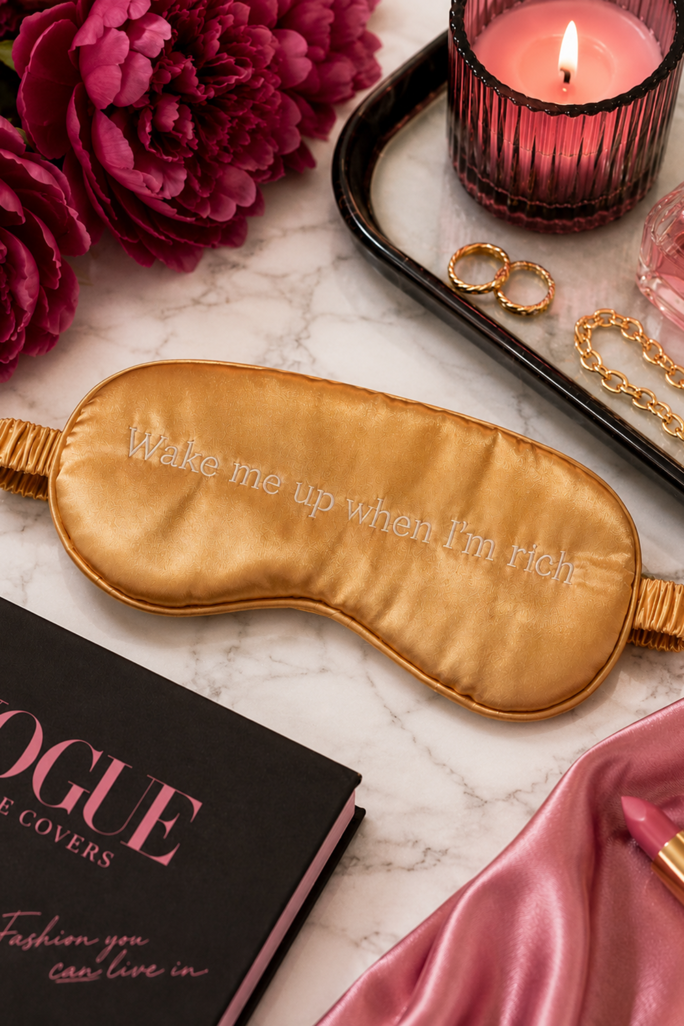 Gold sleep mask with 'Wake me up when I'm rich' text, fashion book, pink flowers, and candle on marble surface